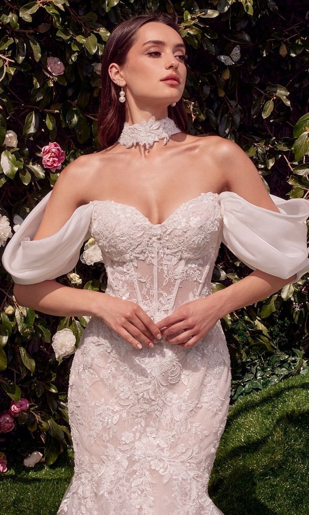 A woman wearing the Andrea and Leo WL031 long wedding dress, a strapless lace mermaid gown with a sweetheart neckline, stands before lush green foliage and pink and white flowers.